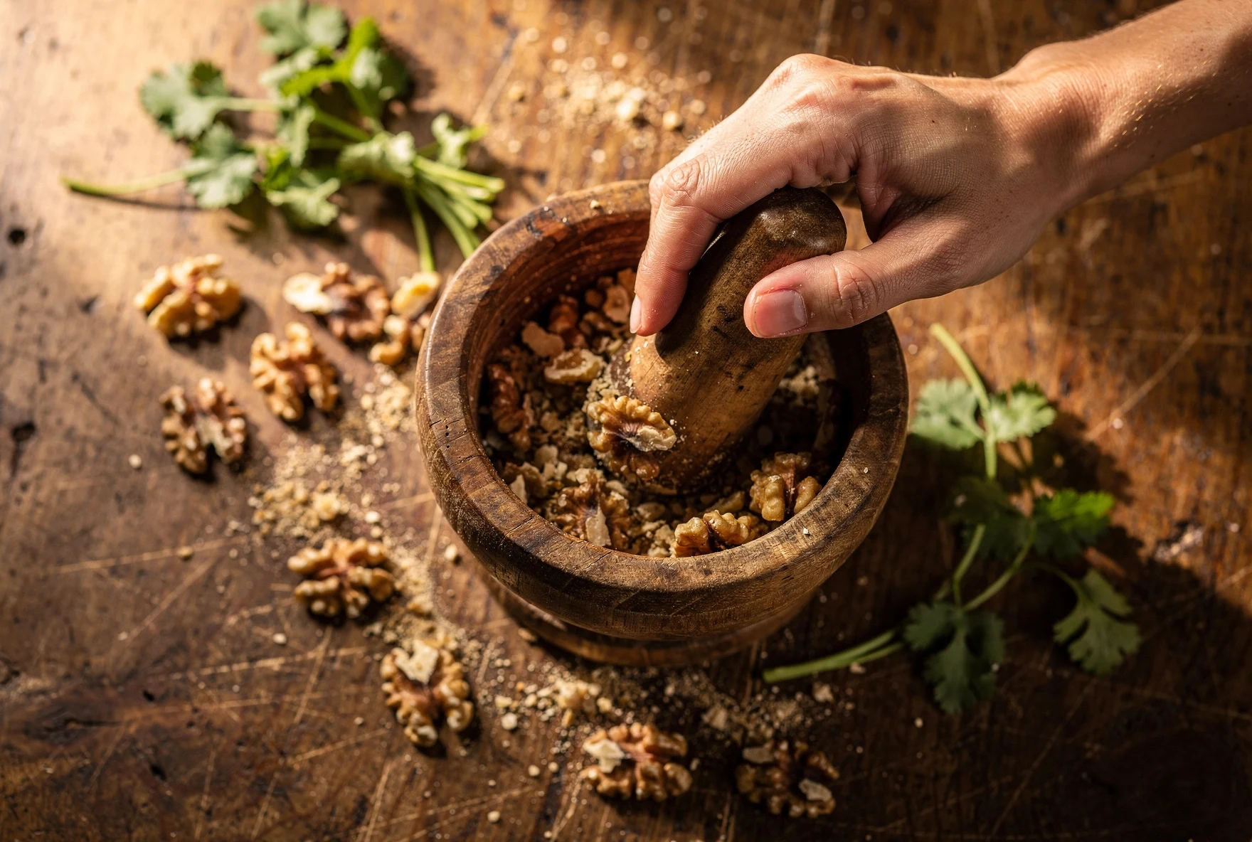 Hands grinding walnuts in a wooden mortar with scattered walnut halves and fresh cilantro on a rustic table