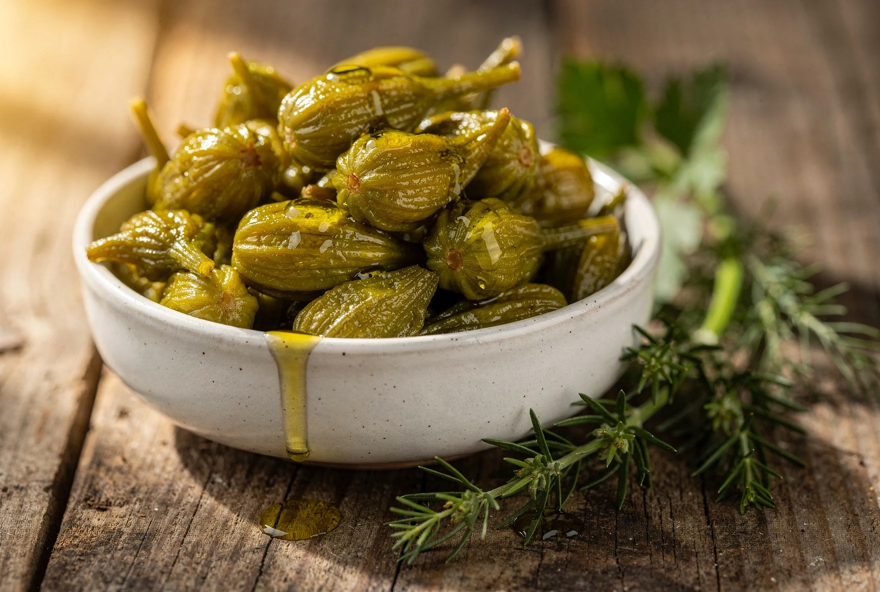 Close-up of pickled jonjoli flower buds in a ceramic bowl with olive oil drizzle and fresh herbs on a wooden table