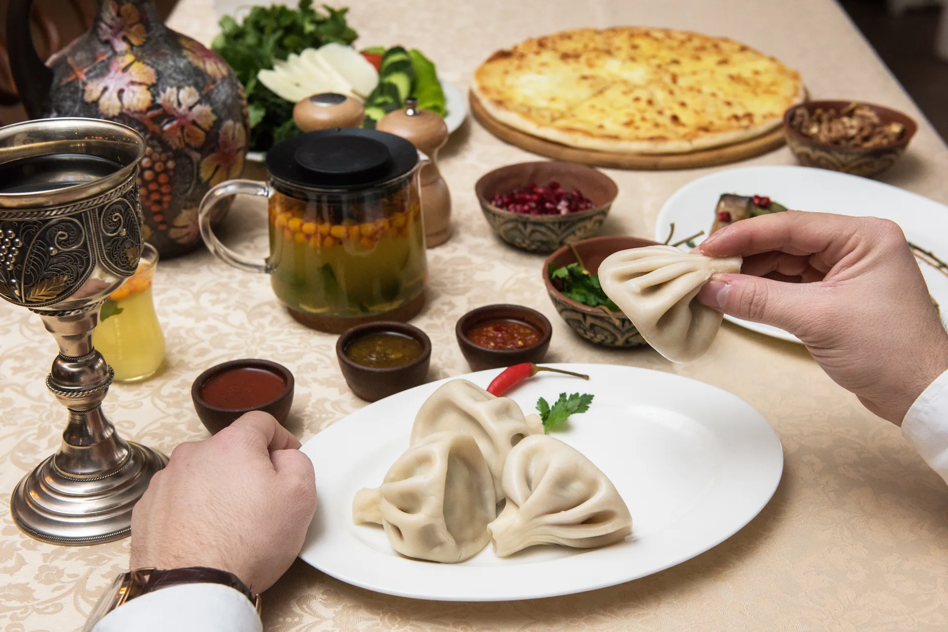 Person holding a khinkali dumpling at a table filled with Georgian dishes and a silver goblet